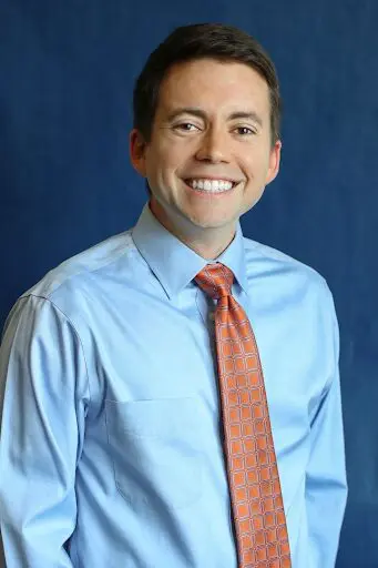 A man in blue shirt and orange tie smiling.